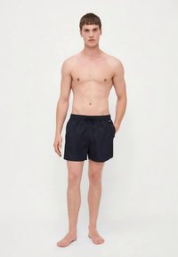 Mlad moški stoji bos, oblečen v črne kopalne hlače z elastiko in vrvico za zategovanje, pred enostavno svetlo ozadje.