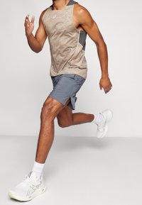 Uomo in canotta beige, pantaloni grigi e scarpe da corsa bianche, catturato a metà corsa su uno sfondo grigio uniforme.