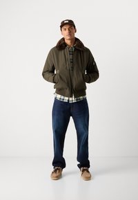 Olivengrøn bomberjakke med pelskrave, ternet skjorte under, mørkeblå jeans og beige mokkasiner. Enkel design, funktionelle lommer.