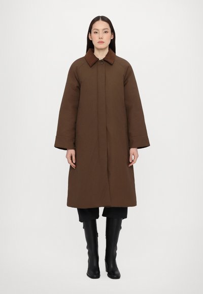 Brown long coat with a structured design, featuring a collared neck, wide sleeves, and a smooth, matte texture. Worn over black pants and boots.