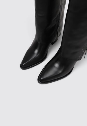 Botas negras de piel con punta estrecha hasta la rodilla y pequeños remaches plateados cerca de la cremallera lateral, mostradas de frente sobre un fondo claro.