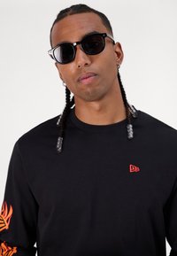 Schwarzes Langarmshirt mit rotem Logo auf der Brust, das orangefarbene Flammenmuster auf dem linken Ärmel aufweist. Schwarze Sonnenbrille rundet den Look ab.