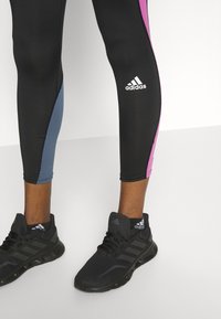 Leggings atletici neri con dettagli rosa e blu, con un logo sul lato. Abbinati a scarpe da corsa nere con texture.