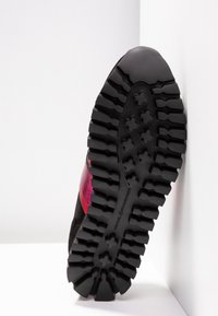 Semelle en caoutchouc noir avec des crampons profonds et des motifs en forme de croix sur une basket avec une bande rouge visible sur le côté.