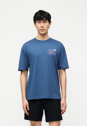 Noor mees tume juustega, kandes sinist Palm Springs Desert Moteli t-särki ja musti lühikesi pükse, seismas ühevärvilise heleda tausta ees.