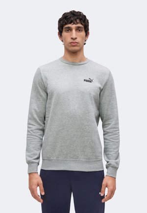 Mann mit dunklen, lockigen Haaren trägt ein hellgraues langärmliges Puma-Sweatshirt und dunkle Hosen, steht vor einem schlichten, hellen Hintergrund.
