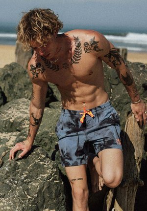 Oberkörperfreier junger Mann mit Tätowierungen und lockigem Haar, der am Strand Felsen erklimmt, trägt blaue Badeshorts mit Palmenmotiven.
