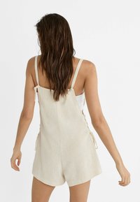 Tuta in lino beige con spalline regolabili e dettagli laterali annodati, caratterizzata da una vestibilità comoda e uno stile corto.