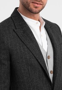 Blazer grigio scuro strutturato con sottili righe a spina, dotato di rever a scalfo e di una singola tasca sul petto. Sotto, un gilet grigio chiaro con bottoni marroni.