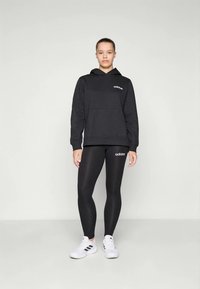 Hoodie preto com bolso frontal e logótipo, combinado com leggings pretas com logótipo. Ténis brancos de desporto com três riscas nas laterais.