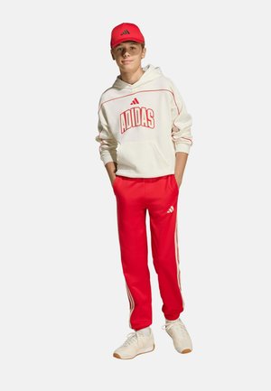 Persona joven con sudadera blanca de Adidas, pantalones deportivos rojos de Adidas, gorra roja y zapatillas blancas, de pie con las manos en los bolsillos.