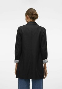Veste noire longue avec un col structuré, des manches trois-quarts et des détails de poignets rayés. Le tissu semble lisse avec une coupe ajustée.