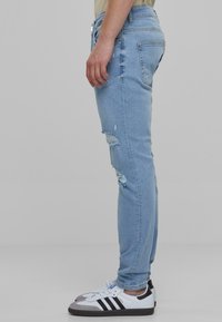 Jeans de mezclilla azul claro con un corte slim, que presentan detalles deshilachados en las rodillas y un diseño estándar de cinco bolsillos, combinados con zapatillas negras y blancas.
