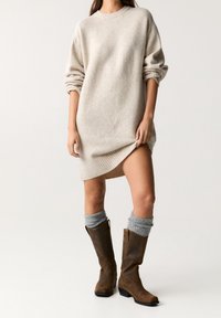 Femme portant une robe pull beige surdimensionnée avec des manches retroussées, des chaussettes grises amples et des bottes en cuir marron jusqu'au mollet sur fond uni.
