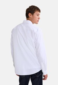 Chemise blanche à manches longues avec un col classique, coupe droite et poignets boutonnés, présentant une texture lisse et un détail de patte de dos.