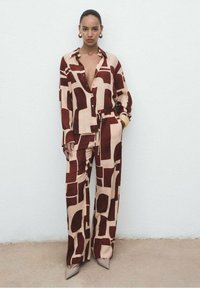 Zweiteiliges Outfit, bestehend aus einem button-up Hemd und weit geschnittenen Hosen. Der Stoff ist mit abstrakten braunen Formen auf einem hellbeigen Hintergrund gemustert.