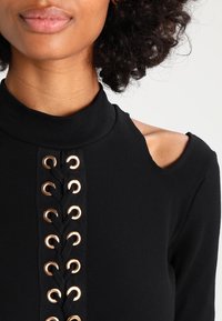 Svart ribbad långärmad topp med en asymmetrisk axelutskärning och en dekorativ snörning framtill med guldfärgade öglor.