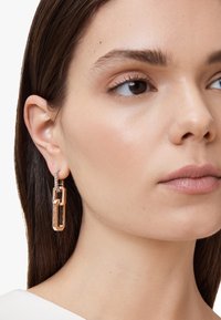 Orecchini pendenti dorati con design geometrico a catena, caratterizzati da una finitura testurizzata e un accento scintillante sulla parte superiore.