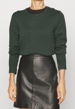 Žena ve tmavě zeleném svetru s dlouhými rukávy a otvory na palce, v černé kožené minisukni, stojící před jednobarevným pozadím.