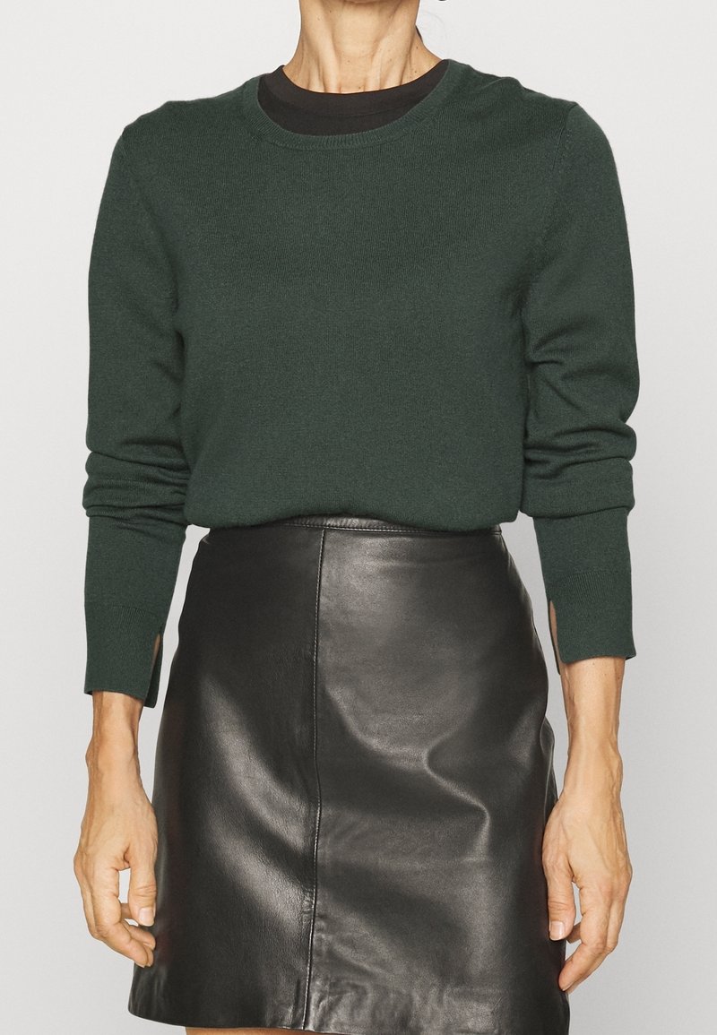 Femme portant un pull à manches longues vert foncé avec des fentes pour les pouces et une mini jupe en cuir noir, debout devant un arrière-plan uni.