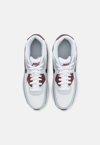 Ténis atléticos brancos e cinzentos com detalhes em bordô, malha e atacadores brancos. Apresenta um colarinho acolchoado e a marca Nike na língua.