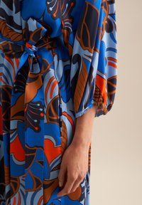Blau gemustertes Kleid mit kräftigen orangen und braunen Blumenmustern, mit einer gebundenen Taille und puffärmel aus einem leichten Stoff.