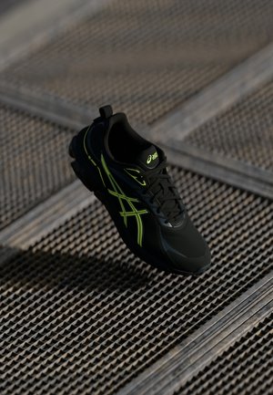 Chaussure de course ASICS noire avec logo et lacets verts néon reposant sur une surface en grille métallique texturée.