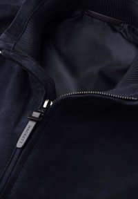 Námořnicky modrá bunda s žebrovaným límcem, stříbrný zip částečně rozepnutý odhalující hladou vnitřní podšívku, jezdec na zipu s raženým nápisem "Hackett".