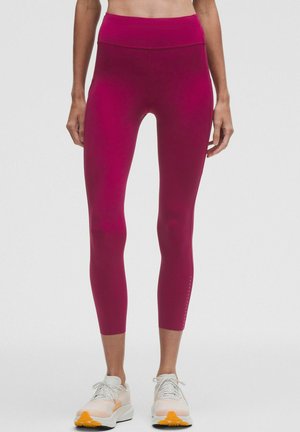 Frau trägt hoch taillierte dunkelrosa kurze Leggings und weiße Sportschuhe und steht vor einem schlichten Hintergrund.