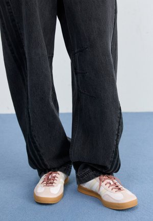 Personne portant un pantalon noir ample et des baskets beiges avec des lacets roses, debout sur un sol bleu face à un mur gris.