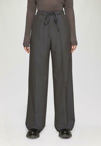 Weite Hosen aus dunkelgrauem Stoff mit einer strukturierten Oberfläche, ausgestattet mit einem elastischen Bund und einem vorderen Bindegürtel. Schwarze Schuhe sind teilweise zu sehen.