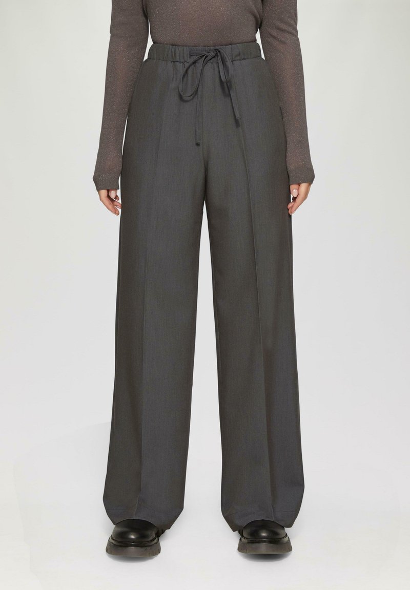 Weite Hosen aus dunkelgrauem Stoff mit einer strukturierten Oberfläche, ausgestattet mit einem elastischen Bund und einem vorderen Bindegürtel. Schwarze Schuhe sind teilweise zu sehen.