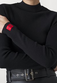Person, der einen schwarzen gerippten Rollkragenpullover mit einem roten Hugo-Logo-Patch am Ärmel und einem schwarzen Ledergürtel mit Metallverschluss trägt.