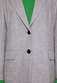 Blazer gris en mélange de laine avec deux boutons noirs, revers crantés et deux poches avant, porté sur un haut côtelé vert.