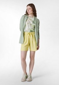 Ljusgrön jacka, top med tie-dye-mönster, gula shorts och beige sneakers med gröna accenter. Mjuka material och avslappnad passform.