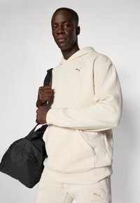 Beige tekstureret hoodie med et lille Puma-logo. Modellen holder en sort træningspose med en lynlås-lomme. Let afslappet pasform.