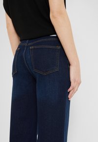 Personne portant un jean denim bleu foncé et un haut noir, vue de dos avec la main reposant sur le côté.