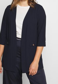 Veste légère bleu marine avec manches aux trois quarts, devant ouvert et deux poches à rabat. Le tissu semble lisse avec une texture subtile.