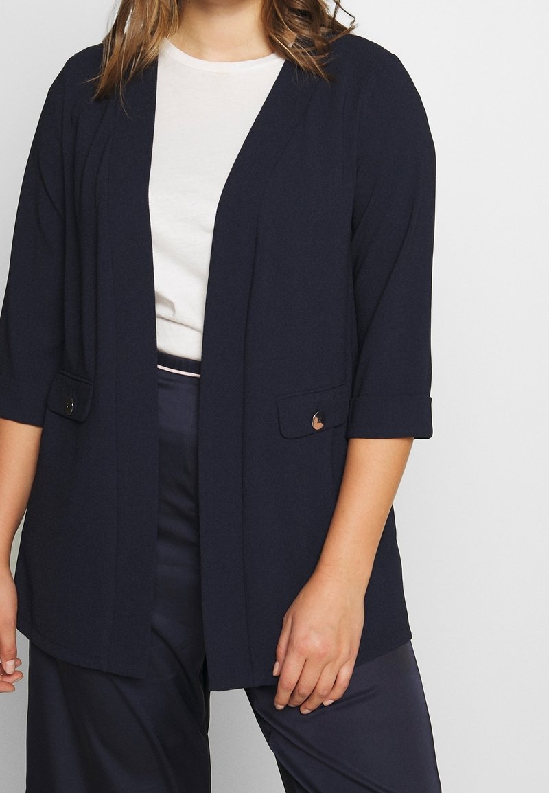 Veste légère bleu marine avec manches aux trois quarts, devant ouvert et deux poches à rabat. Le tissu semble lisse avec une texture subtile.