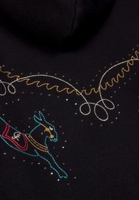 Un tejido negro presenta un bordado colorido de un animal saltando, acentuado con hilos metálicos y detalles decorativos dispersos.