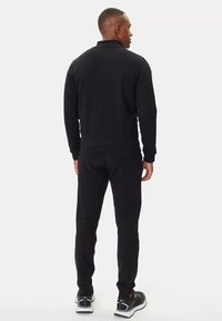 Tuta sportiva nera con colletto zip, polsini a costine e vita con cordino. Tessuto liscio; silhouette aderente. Scarpe da ginnastica visibili sotto.