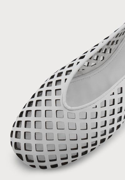 Chaussure plate blanche à bout arrondi présentant un motif en grille de découpes carrées laissant apparaître un matériau intérieur noir.