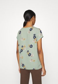 Květinová blůza s krátkým rukávem ve světle zelené barvě, na hladkém materiálu s modrými, šedými a oranžovými květy. Pohled ze zadu ukazuje uvolněný střih a kulatý výstřih.