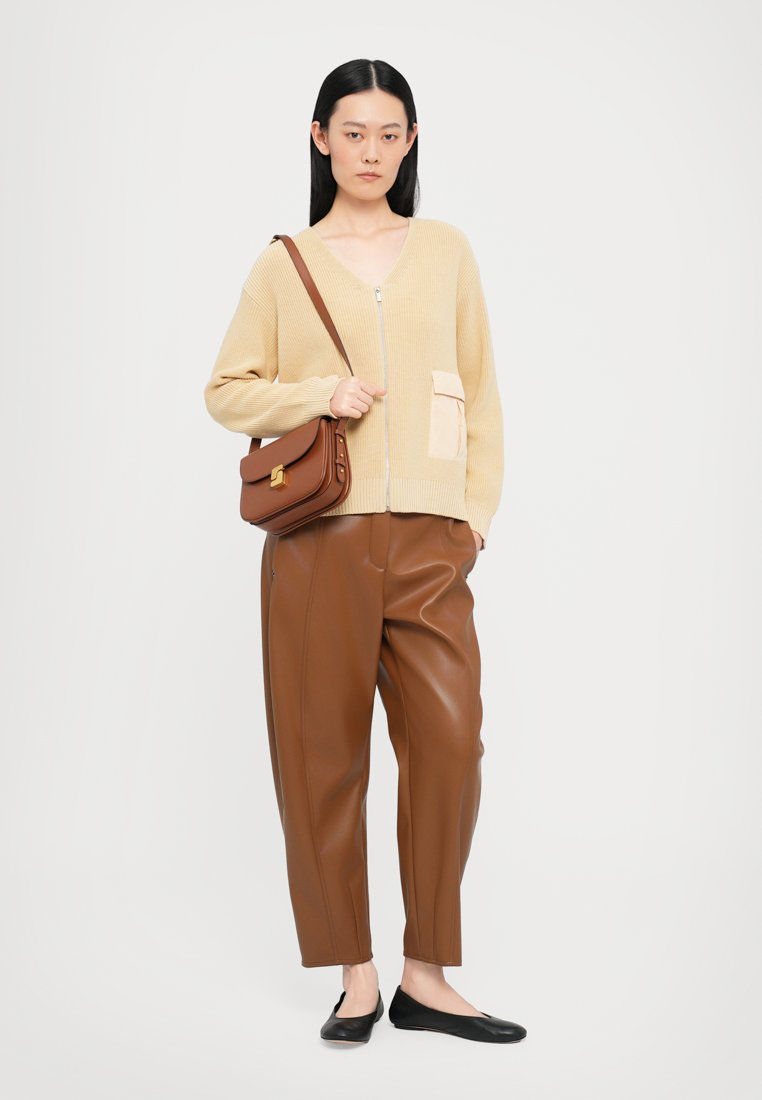 Cardigan en tricot beige avec une fermeture éclair à l'avant, doté d'une poche de couleur claire ; pantalons en faux cuir marron ; ballerines noires ; sac à bandoulière marron.