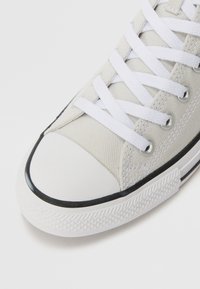 Canvas-Sneaker mit einem hellgrauen Obermaterial, weißen Schnürsenkeln, einer gummierten weißen Zehenkappe und einer strukturierten schwarzen Außensohle mit einem Rautenmuster.