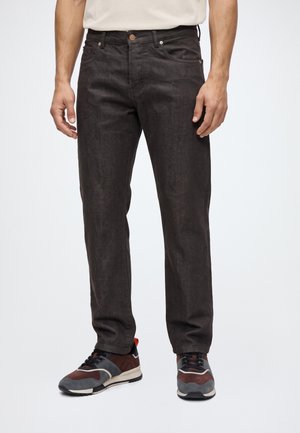 Un mannequin porte un jean noir à jambes droites, une chemise blanche et des baskets bordeaux et grises sur un fond blanc uni.
