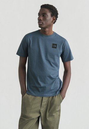 Camicia da uomo a maniche corte in cotone blu scuro, con una toppa con logo nero sul petto sinistro, abbinata a pantaloni verde oliva.