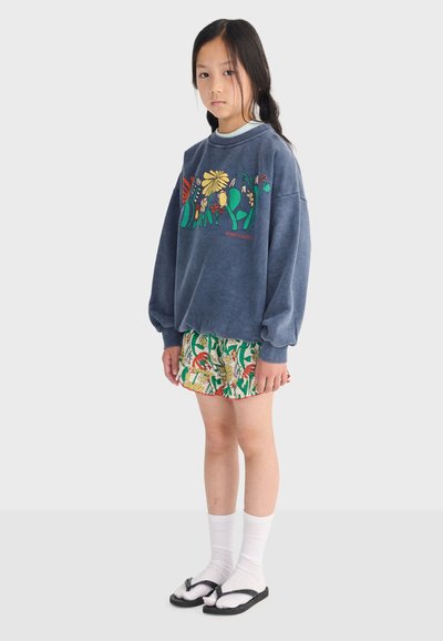 Chica con cabello largo y oscuro que lleva una sudadera azul con estampado floral, pantalones cortos con patrón, calcetas blancas y sandalias negras, de pie frente a un fondo liso.