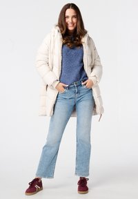 Lichtbeige pufferjack met een bontgevoerde capuchon, getextureerde blauwe trui en lichtblauwe recht gesneden jeans. Borden sneakers met witte accenten.