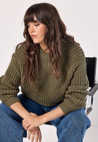 Chunky olijfgroene gebreide trui met een gestructureerd golfpatroon, gecombineerd met blauwe denim jeans met wijde pijpen. Model zit op een zwarte stoel.